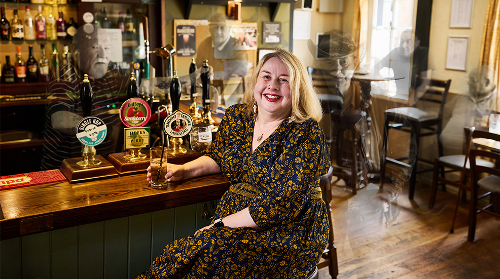 Nat Doig sat in an English pub with a drink, smiling at the camera, with ghosts edited behind her
